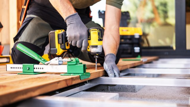 A contractor using a power drill to fasten wooden deck boards on a frame while building a custom outdoor deck.