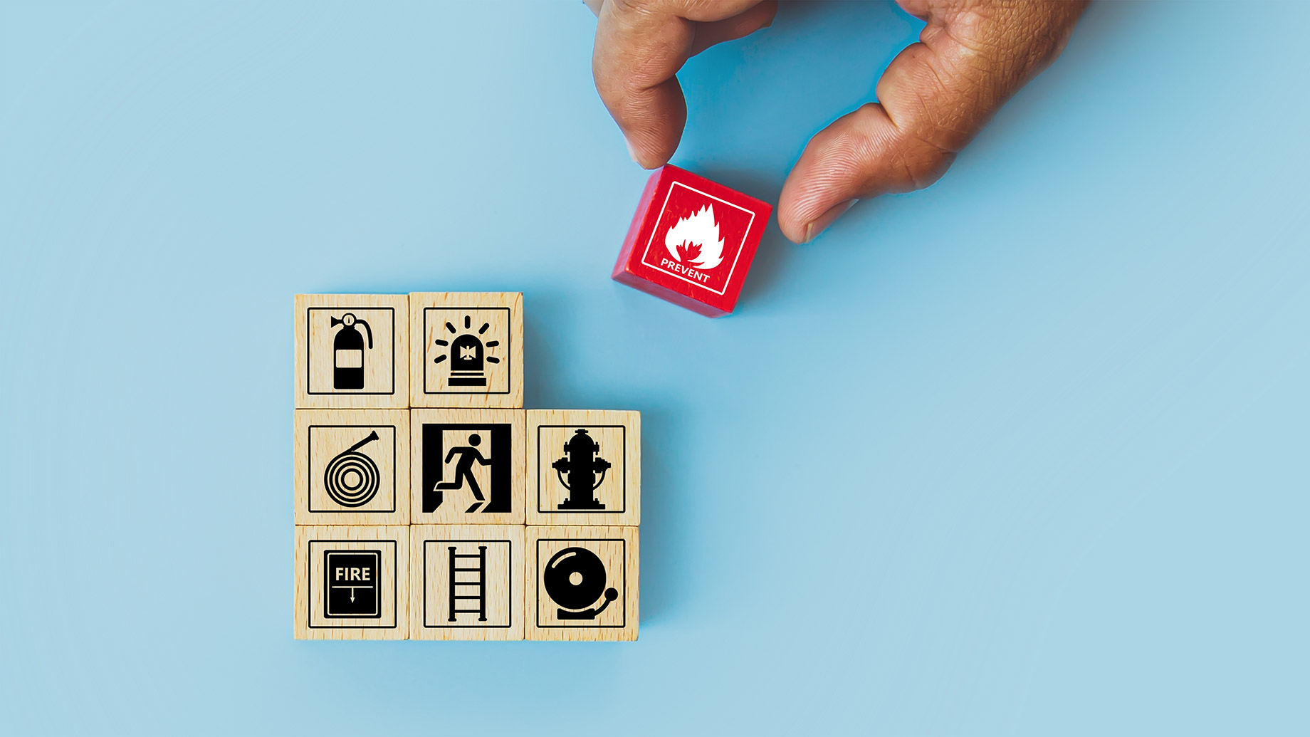 Hand placing a red fire prevention block above wooden cubes showing fire safety icons including extinguisher, alarm, hose, hydrant, escape route, ladder, and alarm bell.