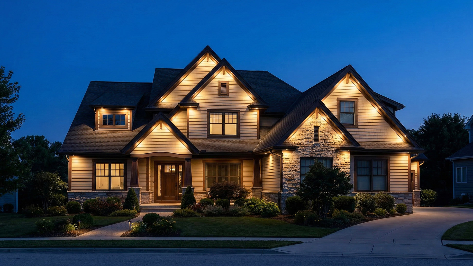 A nighttime view of a large, two-story custom home featuring beige siding, stone veneer accents, and multiple gables. The structure is dramatically illuminated by warm uplighting and soffit lights under a dark blue sky.