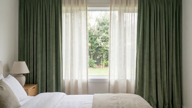 A bedroom window dressed with heavy sage green velvet curtains layered over sheer white drapes. Sunlight filters through the sheers from a green yard outside. A smartly made bed with white linens and a beige throw sits in the foreground, next to a wooden nightstand and a ceramic lamp.