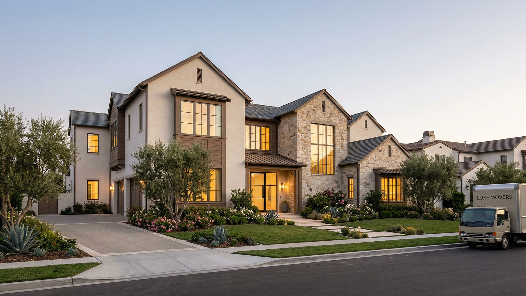 A large two-story luxury house with a stucco and stone exterior, manicured landscaping, and a white moving truck parked on the street out front.