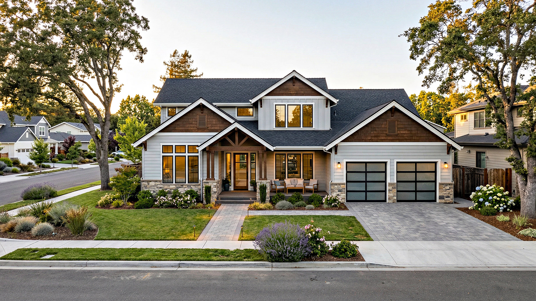 A two-story modern craftsman house with gray siding, wood accents, and a stone foundation, set on a manicured lawn with a paver driveway, lush landscaping, and a two-car garage.