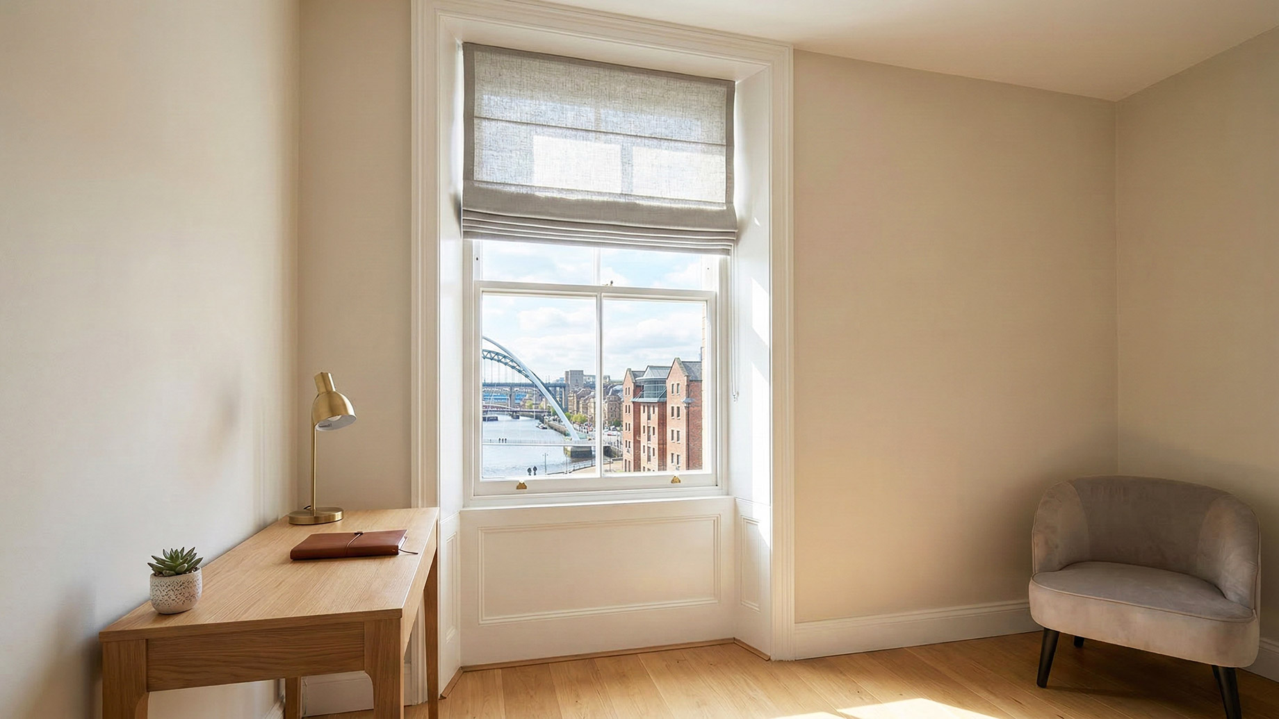An interior of a bright home office with a wooden desk and lamp near a window. Outside, the Newcastle Tyne Bridge is visible over water. A grey velvet armchair sits in the far corner.