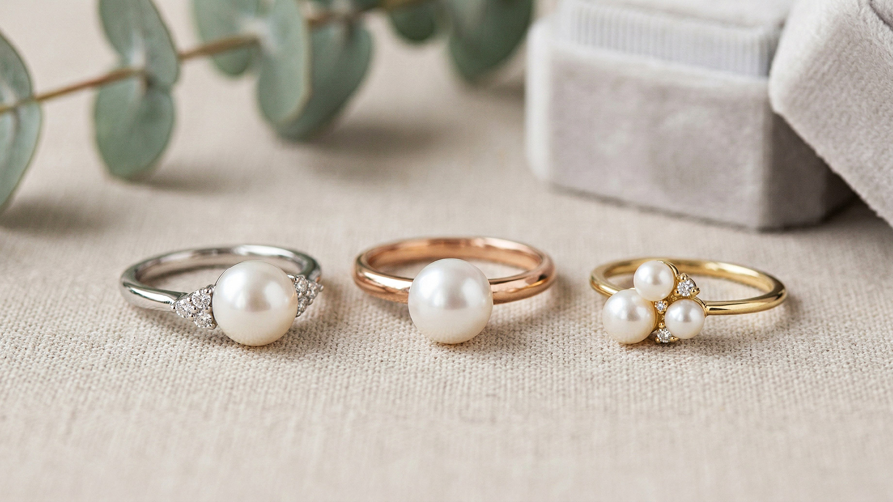 A macro shot from left to right showing three different pearl rings on a cream-colored textured cloth: a white gold ring with a single pearl and diamond accents; a rose gold ring with a single solitaire pearl; and a yellow gold ring with a cluster of three pearls and diamonds. In the softly blurred background are a grey velvet box and eucalyptus leaves.