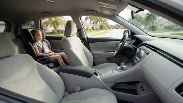 An interior view of a car showing a child safely secured in a forward-facing car seat in the back row, with the front passenger seat left completely empty. A sunny residential street is visible through the vehicle's windows.