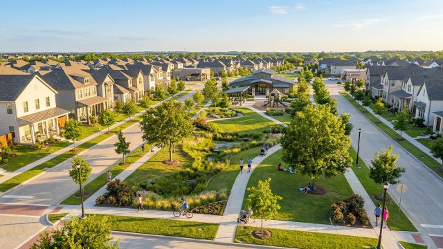 An aerial view of a modern neighborhood featuring contemporary homes built around a large, landscaped central park where residents are walking, cycling, and relaxing.