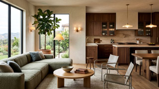An open-concept living space featuring a sage green sectional sofa, a round wooden coffee table, and two modern metal-framed accent chairs. A large indoor tree stands next to floor-to-ceiling windows. In the background, a kitchen displays reeded wood cabinets, stone countertops, and warm pendant lighting beside a solid wood dining table with upholstered chairs.