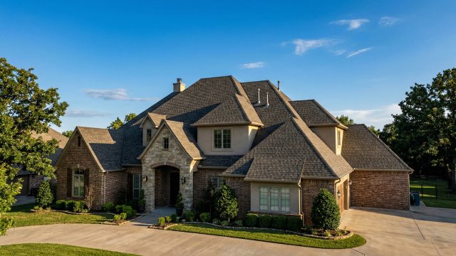 A large, luxury brick and stone house under a clear blue sky, featuring a complex, beautifully maintained multi-gabled shingle roof. The home is surrounded by green trees, a manicured lawn, and a curved concrete driveway.
