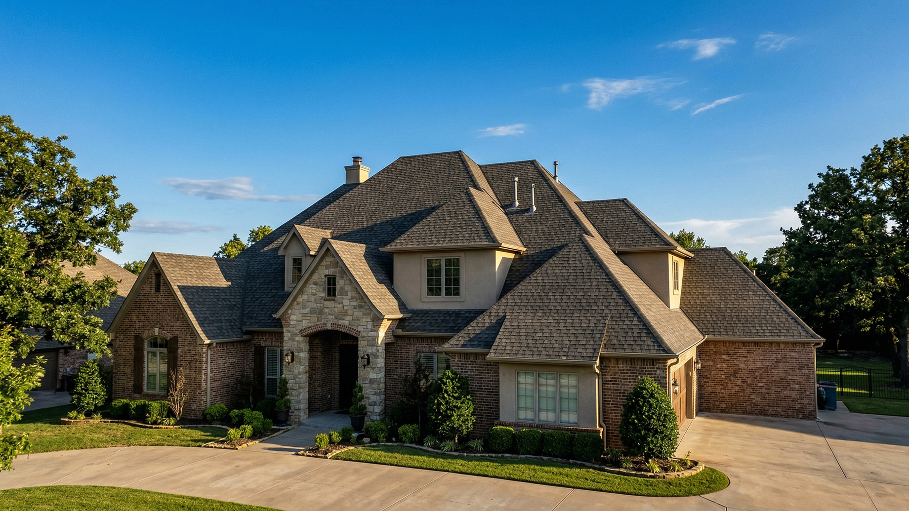 A large, luxury brick and stone house under a clear blue sky, featuring a complex, beautifully maintained multi-gabled shingle roof. The home is surrounded by green trees, a manicured lawn, and a curved concrete driveway.