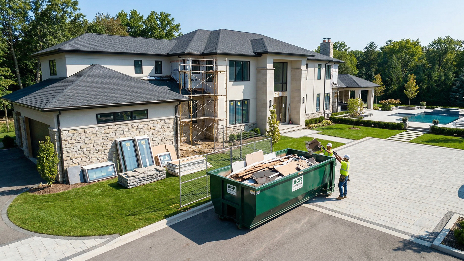 A large green ACR roll-off dumpster sits on the expansive paved driveway of a luxury home undergoing renovations. Two construction workers wearing hard hats and high-visibility vests are actively tossing demolition debris into the container. Scaffolding and neatly organized building materials are visible near the house.