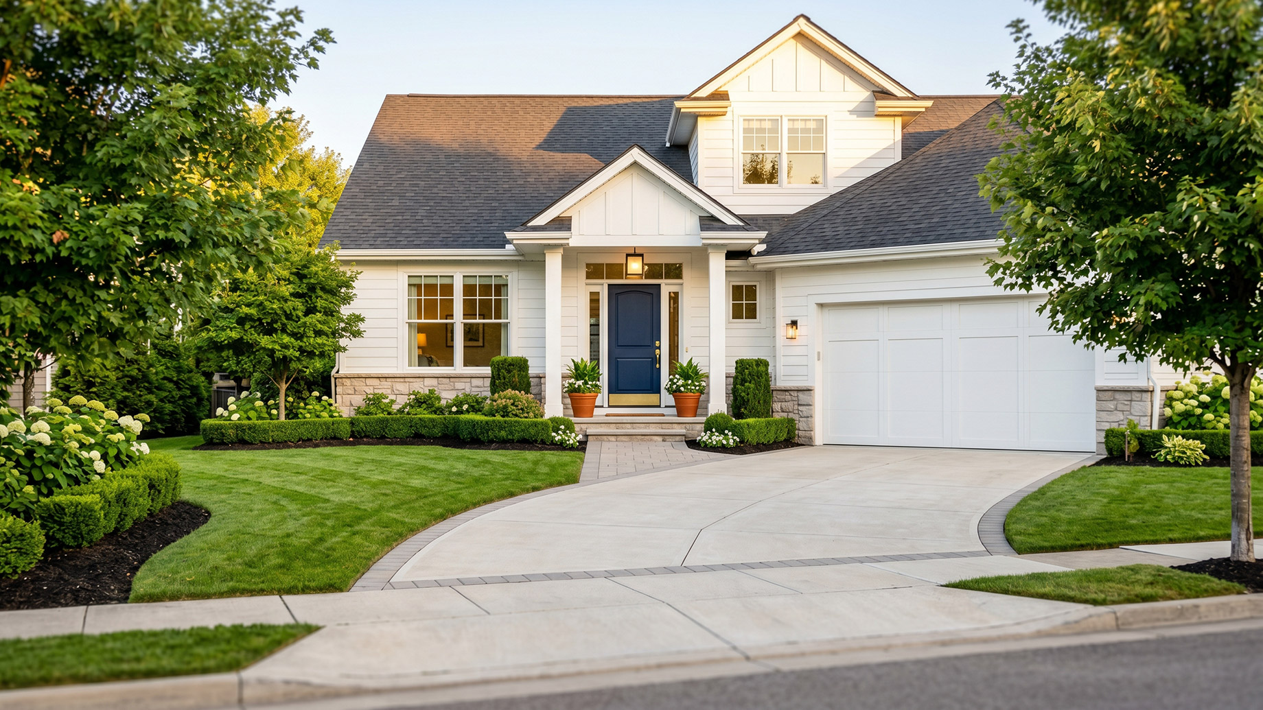 An elegant, two-story white house with stone accents, a deep blue front door, and tidy green landscaping including bushes and flowering hydrangeas, all under a warm dusk sky. A concrete driveway and paver path lead to the entrance.