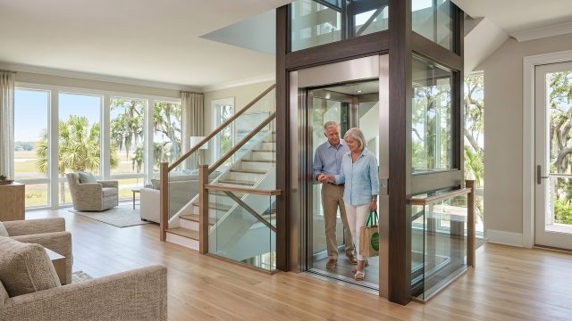 An older couple in coastal clothes steps from a modern glass elevator into a luxurious Lowcountry home, next to a staircase and large windows with a coastal view.