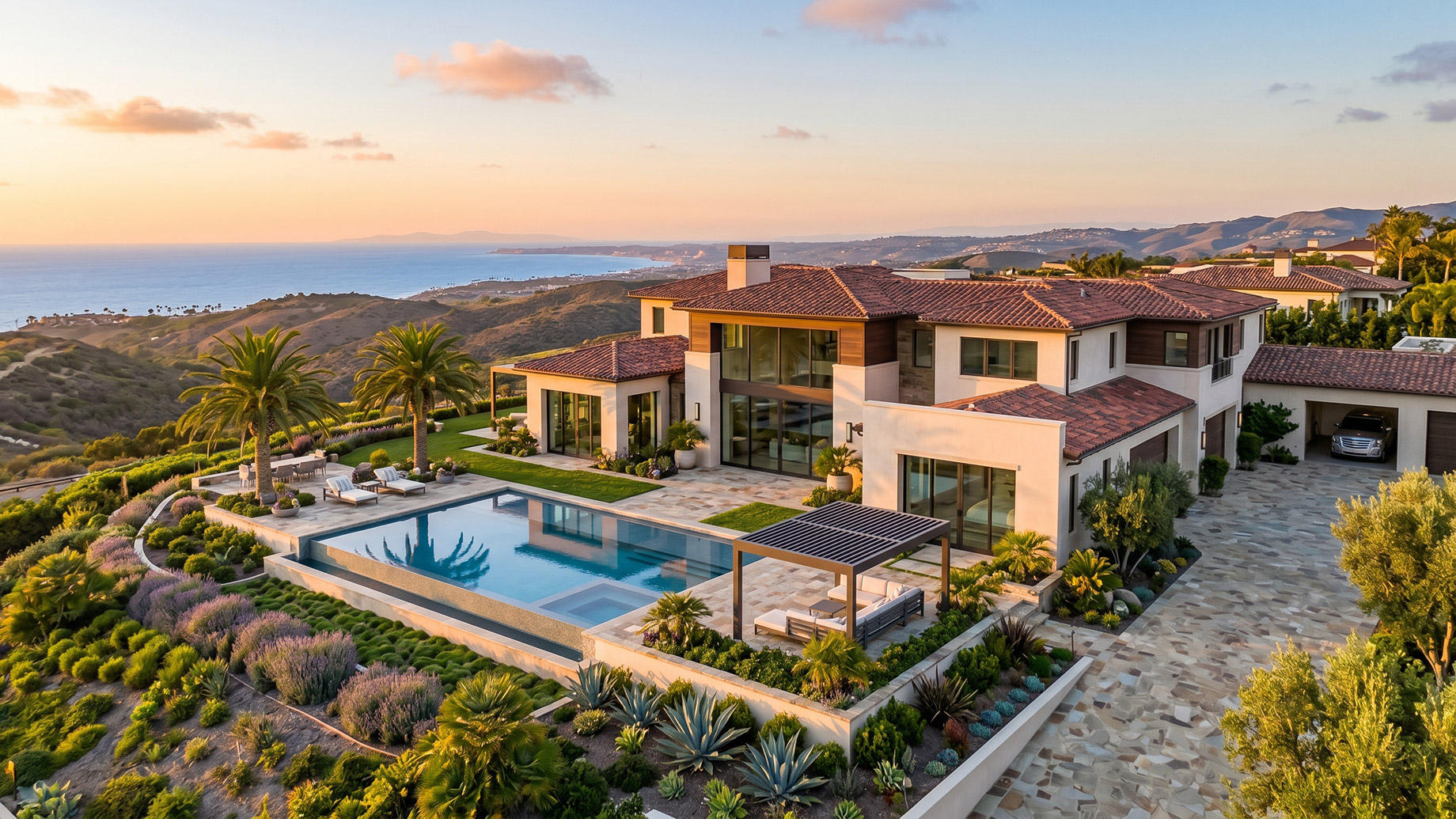 An aerial view of a sprawling luxury mansion perched on a hillside at sunset. The multi-level home features white stucco walls, a terracotta tile roof, and large glass windows. A large swimming pool and a modern pergola sit on a stone patio overlooking the ocean and distant coastline. The property is surrounded by manicured landscaping, including palm trees, lavender, and native coastal plants.