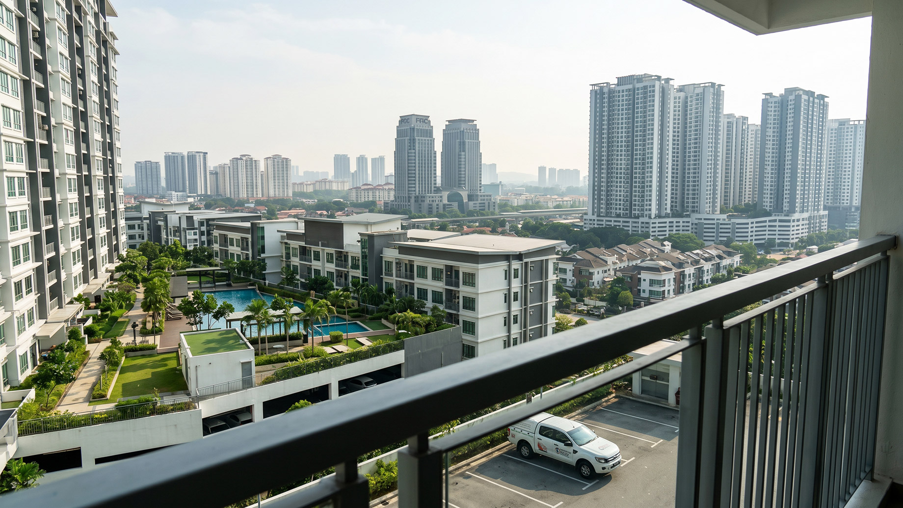 A view from an apartment balcony looking out over modern condominium complexes and high-rise buildings in Puchong. Below, a white service truck is parked near a communal swimming pool and landscaped gardens.