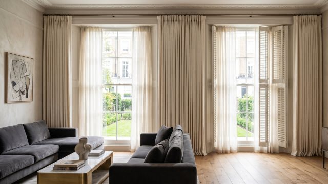 A bright, luxurious living room featuring tall sash windows dressed with soft, floor-length sheer curtains and solid white plantation shutters. The natural light illuminates a dark grey velvet sofa, hardwood floors, and a marble coffee table.