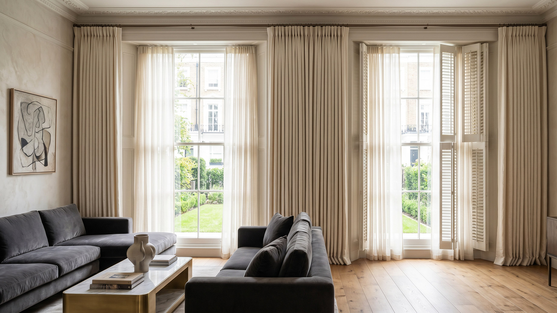 A bright, luxurious living room featuring tall sash windows dressed with soft, floor-length sheer curtains and solid white plantation shutters. The natural light illuminates a dark grey velvet sofa, hardwood floors, and a marble coffee table.