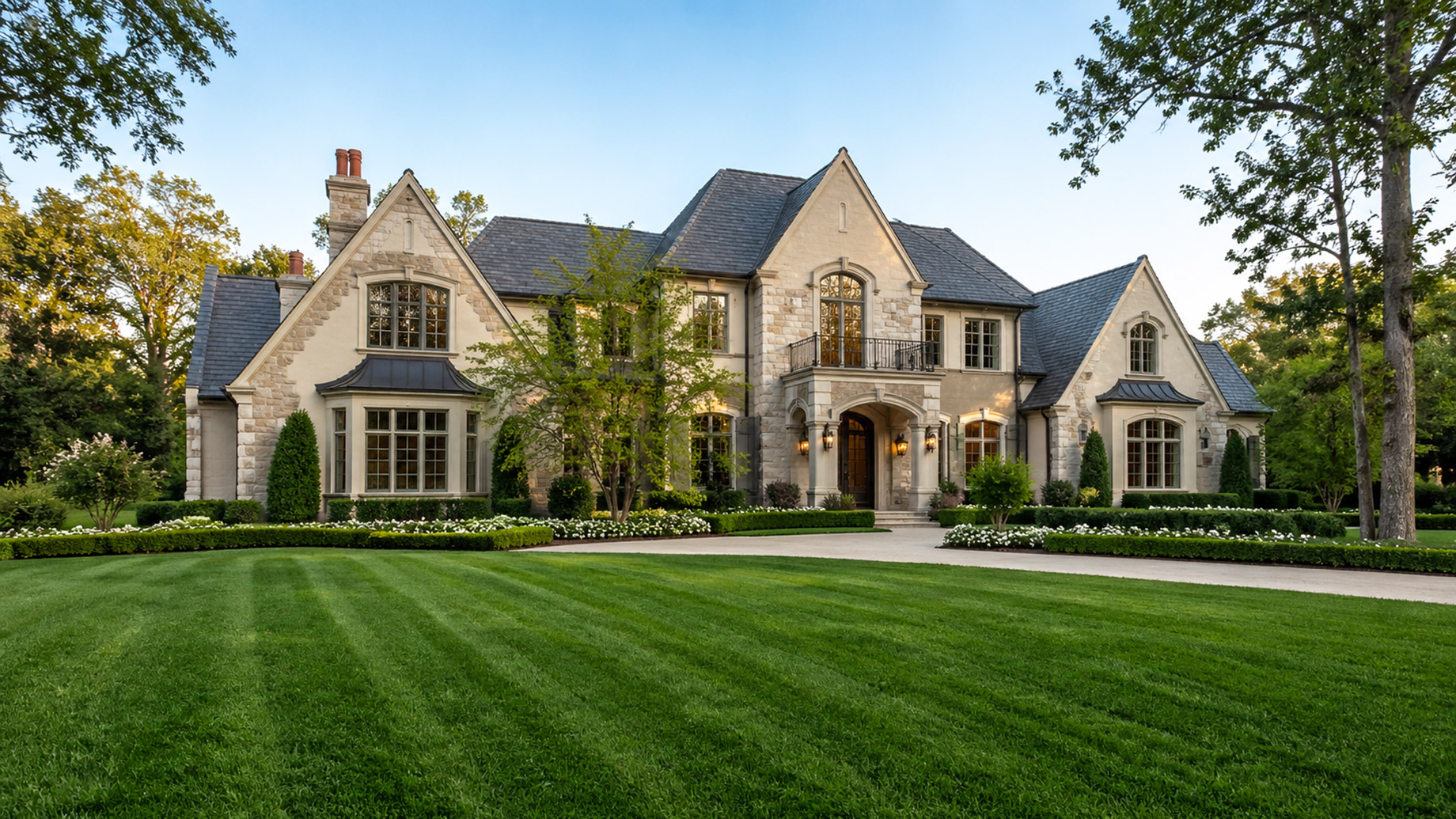 Elegant luxury house with a lush green manicured front lawn, trimmed hedges, and landscaped gardens in soft daylight.