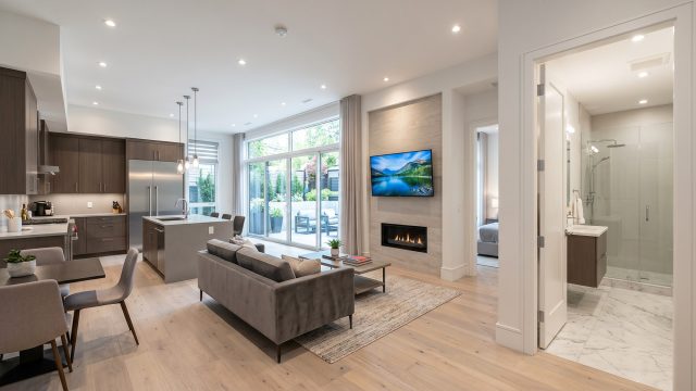 A brightly lit, fully renovated modern basement apartment featuring a full kitchen with an island, a living area with a built-in fireplace and TV, a luxurious glass-door bathroom, and large sliding glass doors opening to a sunken outdoor patio.