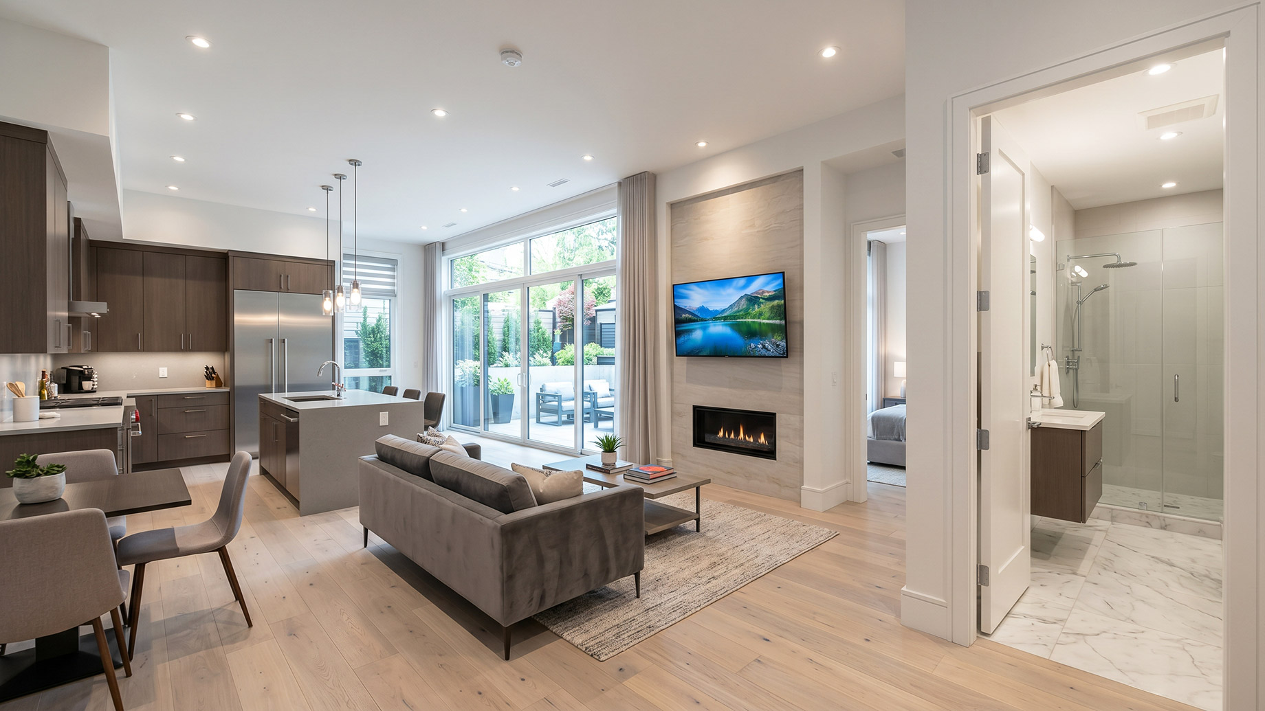 A brightly lit, fully renovated modern basement apartment featuring a full kitchen with an island, a living area with a built-in fireplace and TV, a luxurious glass-door bathroom, and large sliding glass doors opening to a sunken outdoor patio.