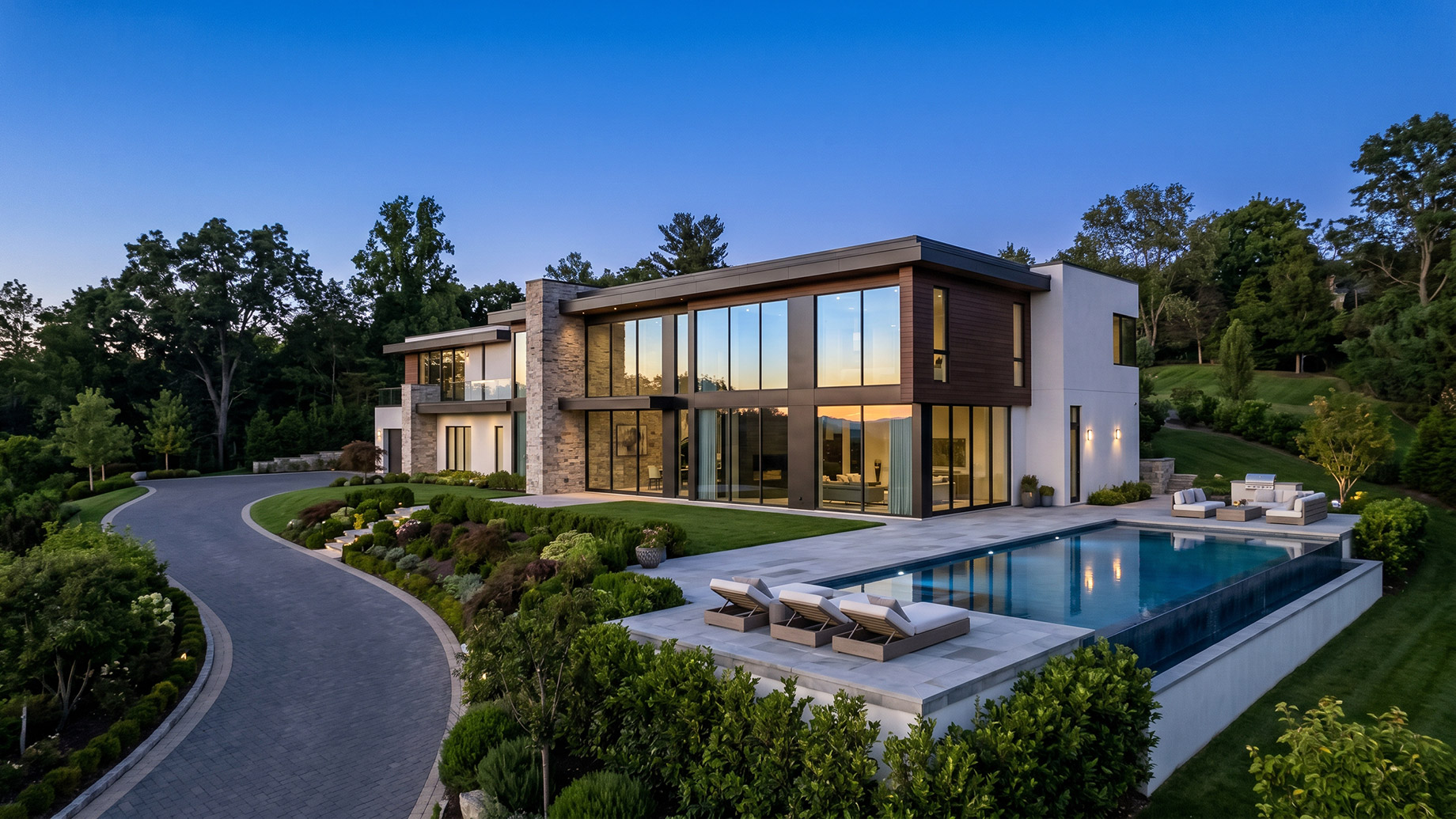 A striking modern luxury estate at dusk, featuring expansive glass windows glowing from within, a pristine infinity pool with lounge chairs, and a sweeping paved driveway surrounded by lush, manicured landscaping.