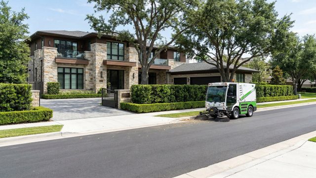 A compact, white and green street sweeping vehicle cleaning the edge of a paved road in front of a large luxury home with manicured hedges and mature trees on a bright, sunny day.