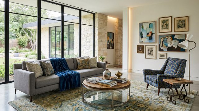 An inviting modern living room featuring a gray sofa with a blue throw, a patterned rug, a geometric armchair, a gallery wall of art, and large black-framed windows looking out onto a lush backyard.