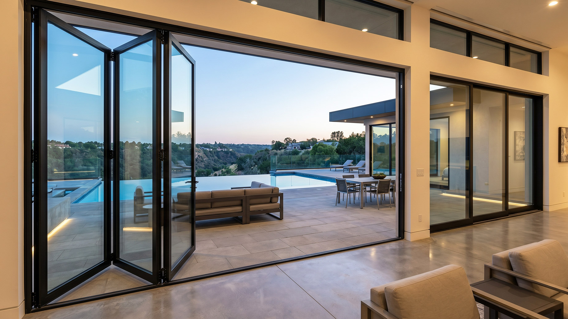 A modern luxury living room featuring large, matte black-framed bi-fold glass doors opened wide, revealing a seamless transition to a spacious outdoor patio with lounge furniture, an infinity pool, and a scenic view of rolling hills at twilight.