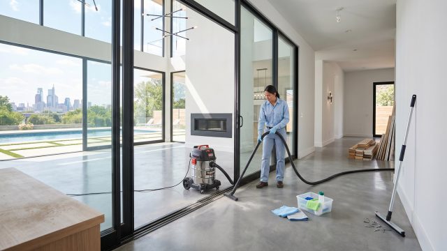 A woman in work clothes vacuums the track of a modern sliding glass door in a house with a concrete floor. Beyond the large windows, a city skyline and pool are visible. Cleaning supplies and construction lumber are on the floor.