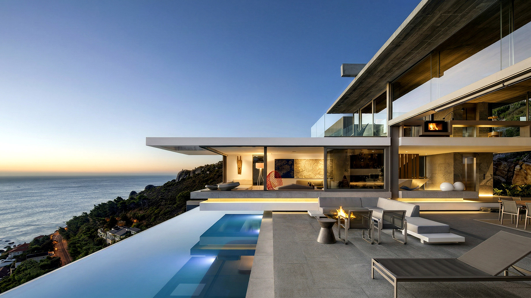 A modern, multi-story luxury villa built into a coastal hillside at dusk, featuring an illuminated infinity pool, an outdoor patio with a modern fire pit, and expansive floor-to-ceiling windows glowing with warm interior light overlooking the ocean.
