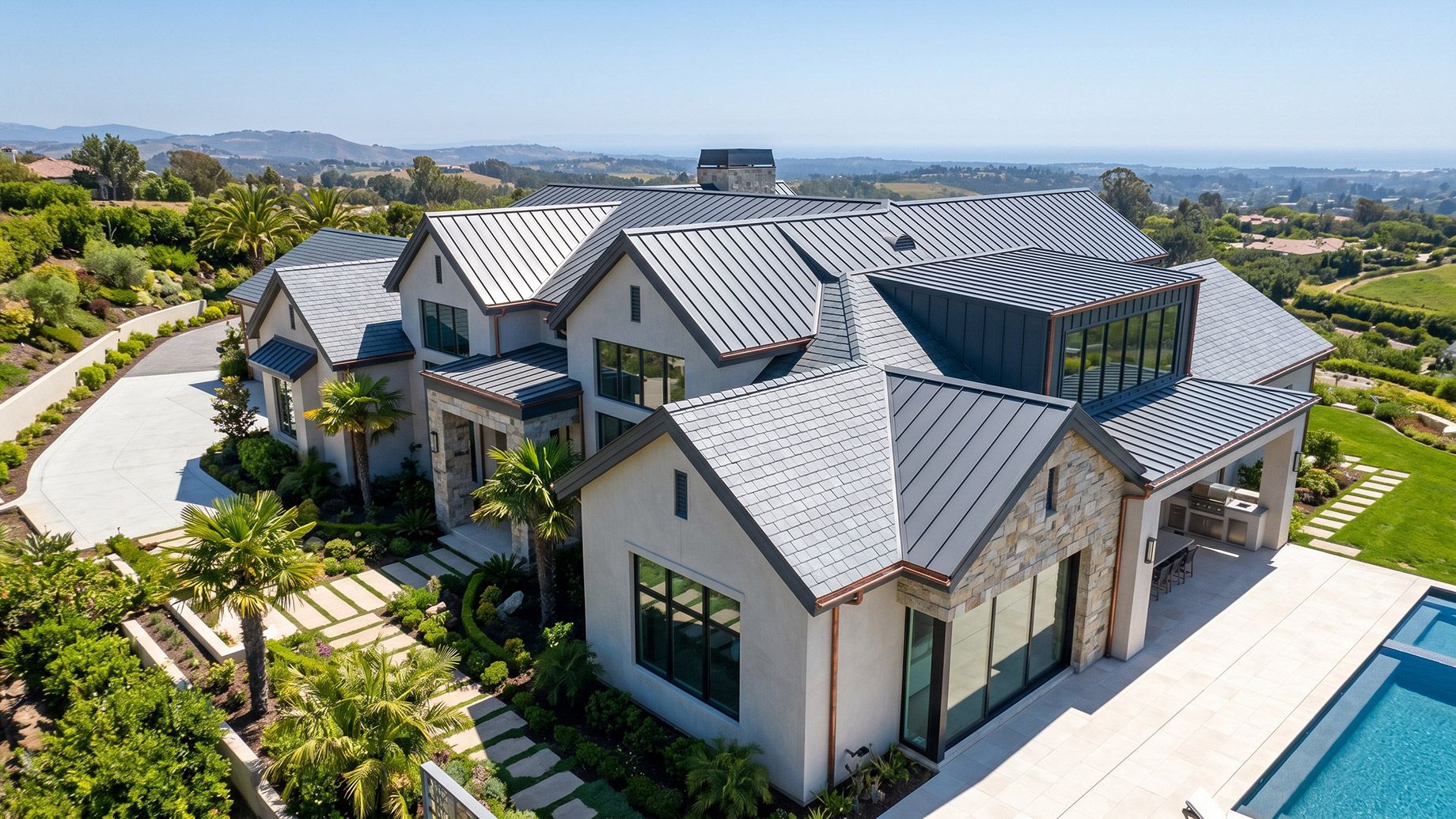 Aerial view of a sprawling luxury home with white stucco walls, stone accents, and a large, uniform dark grey metal roof, surrounded by lush landscaping, palm trees, and a swimming pool.