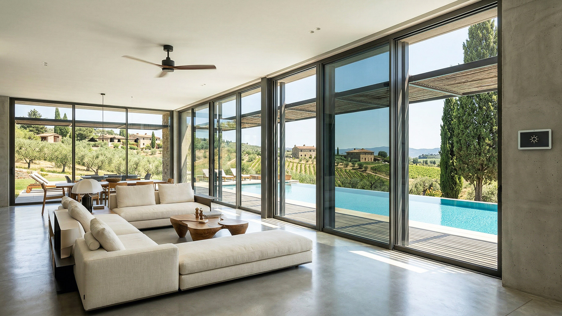 A bright, modern living room featuring a large white sectional sofa and polished concrete floors. Expansive floor-to-ceiling tinted windows look out onto an outdoor infinity pool and a sun-drenched Tuscan landscape. A modern smart thermostat is mounted on a concrete wall on the right side of the room.