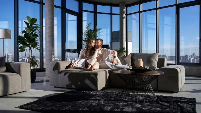 A couple relaxing comfortably together on a large, plush grey sectional sofa in a modern, high-rise apartment. The spacious room features floor-to-ceiling windows offering a panoramic city view, abundant natural light, a minimalist round coffee table, and tall potted indoor plants.