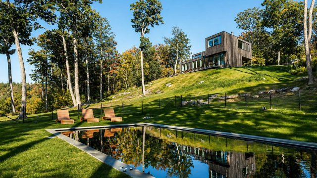 Modern dark-wood house on a grassy hillside overlooking a reflective pool, surrounded by tall trees in a quiet Hudson Valley-style landscape.