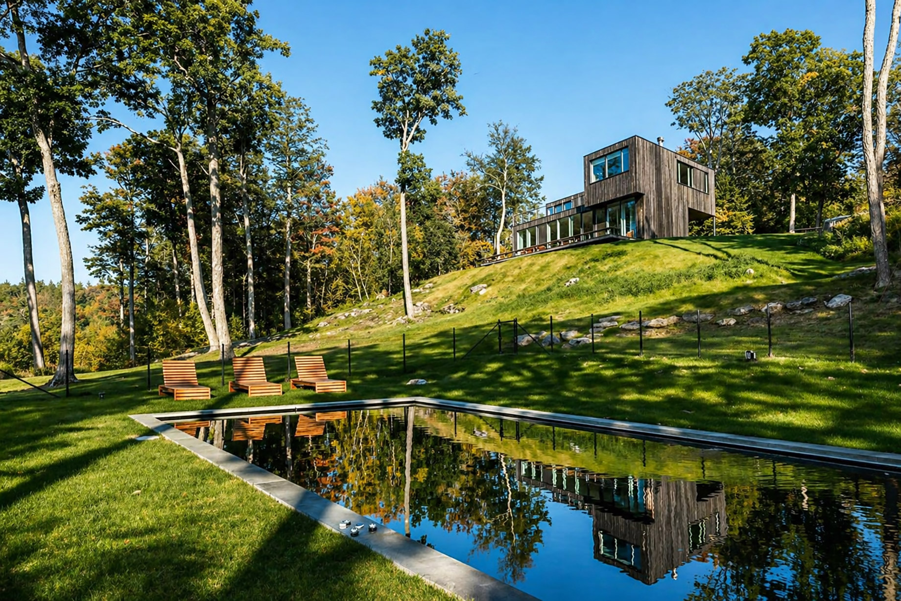 Modern dark-wood house on a grassy hillside overlooking a reflective pool, surrounded by tall trees in a quiet Hudson Valley-style landscape.