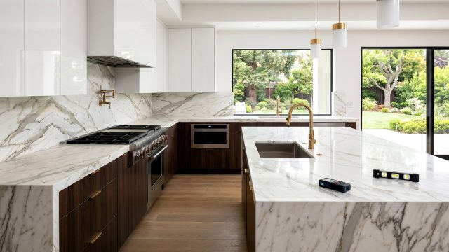 A bright, high-end kitchen featuring custom dark wood cabinets and a large white marble island with gold fixtures. A laser measuring tool and a spirit level rest on the flawless countertop, highlighting the precision of professional installation.