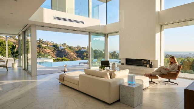 A bright, modern luxury living room with high ceilings, white marble floors, and floor-to-ceiling windows opening to a patio with a swimming pool and a hillside view. A woman relaxes in a leather lounge chair reading a book. The pristine space features a sleek linear air vent high on the wall and a small air purifier, emphasizing clean indoor air.
