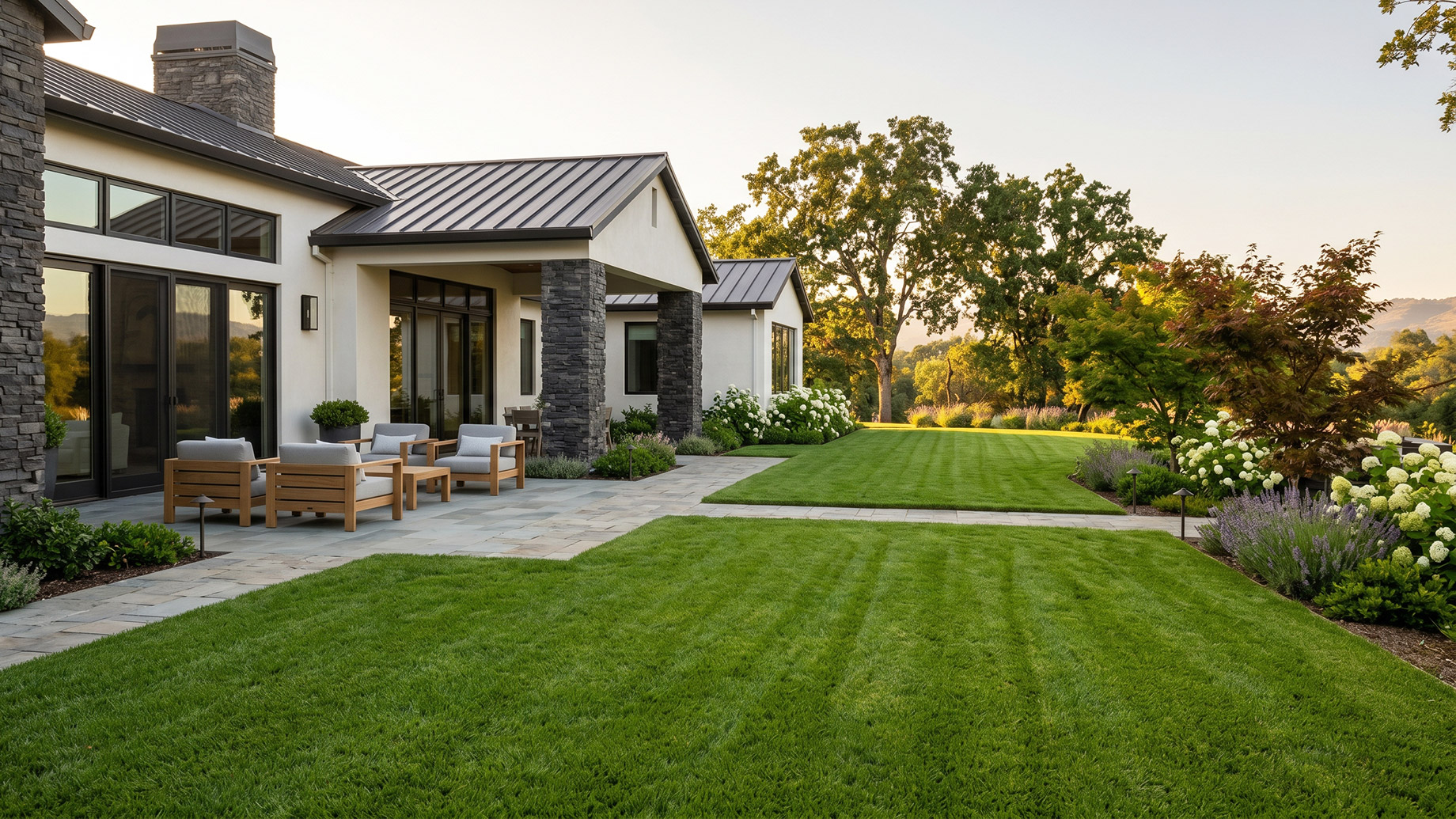 A lush, perfectly manicured green lawn with clean mowing stripes in the backyard of a modern luxury home. The yard features a stone patio with wooden lounge chairs, well-tended garden beds with white flowers, and mature trees in the background under soft sunlight.