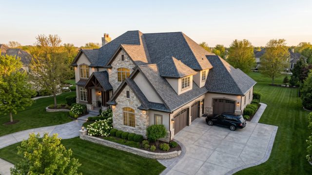 Aerial view of a large luxury estate with a stone and stucco facade, complex gabled roof, and three-car garage. A black SUV is parked on the paved driveway amidst manicured lawns, hydrangeas, and trees under warm evening light.
