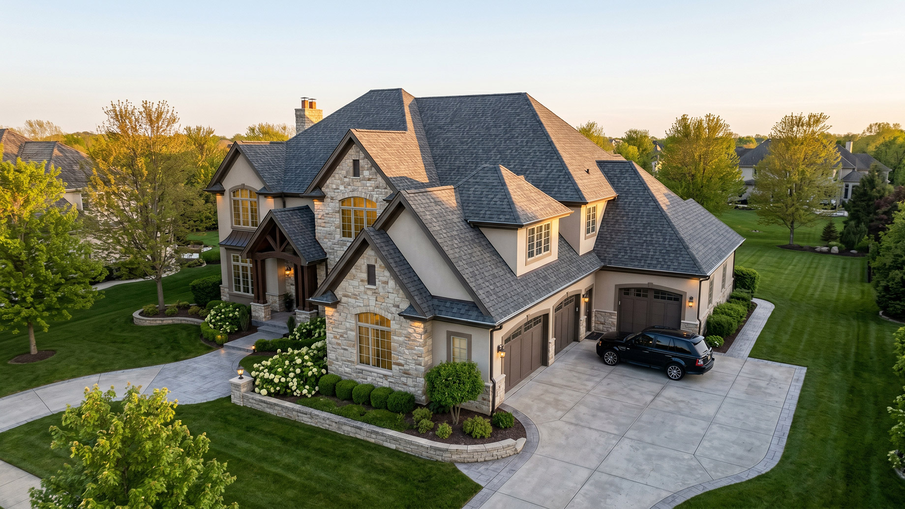 Aerial view of a large luxury estate with a stone and stucco facade, complex gabled roof, and three-car garage. A black SUV is parked on the paved driveway amidst manicured lawns, hydrangeas, and trees under warm evening light.