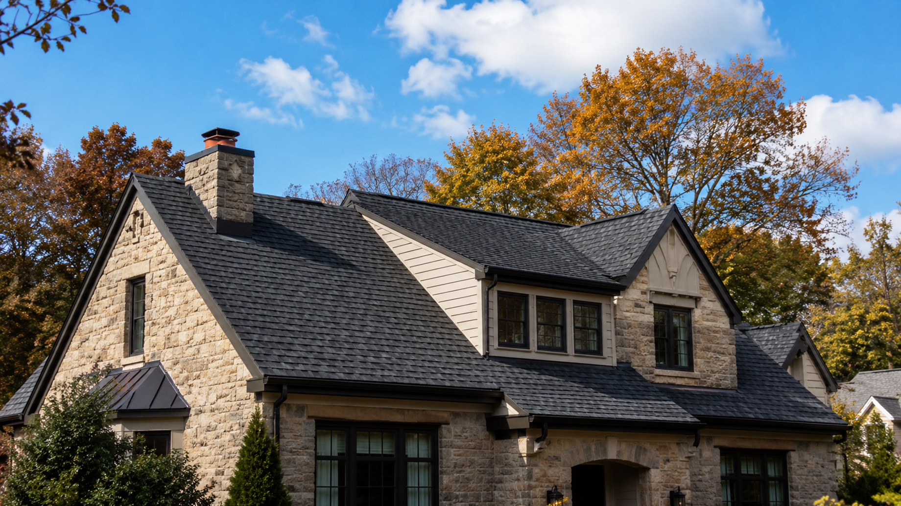 Stone Pittsburgh home with a dark shingle roof, black gutters, chimney, autumn trees, blue sky, and scattered clouds.
