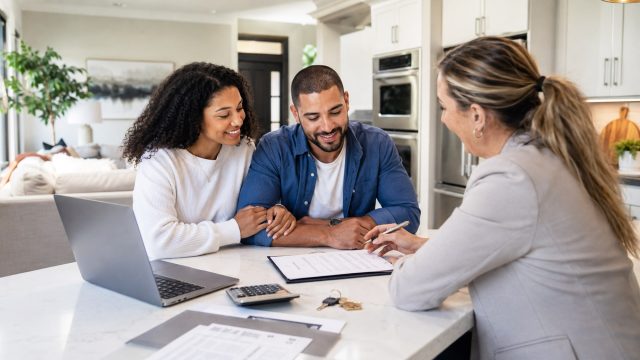 First-time home buyers meeting with a mortgage advisor in a modern kitchen while reviewing loan paperwork, with keys, laptop, and calculator on the counter.