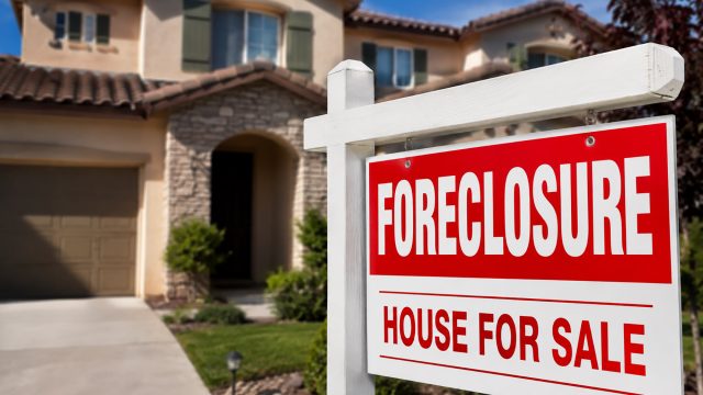 Red and white foreclosure house for sale sign in front of a suburban California home with tan stucco, tile roofing, landscaped yard, and bright blue sky.