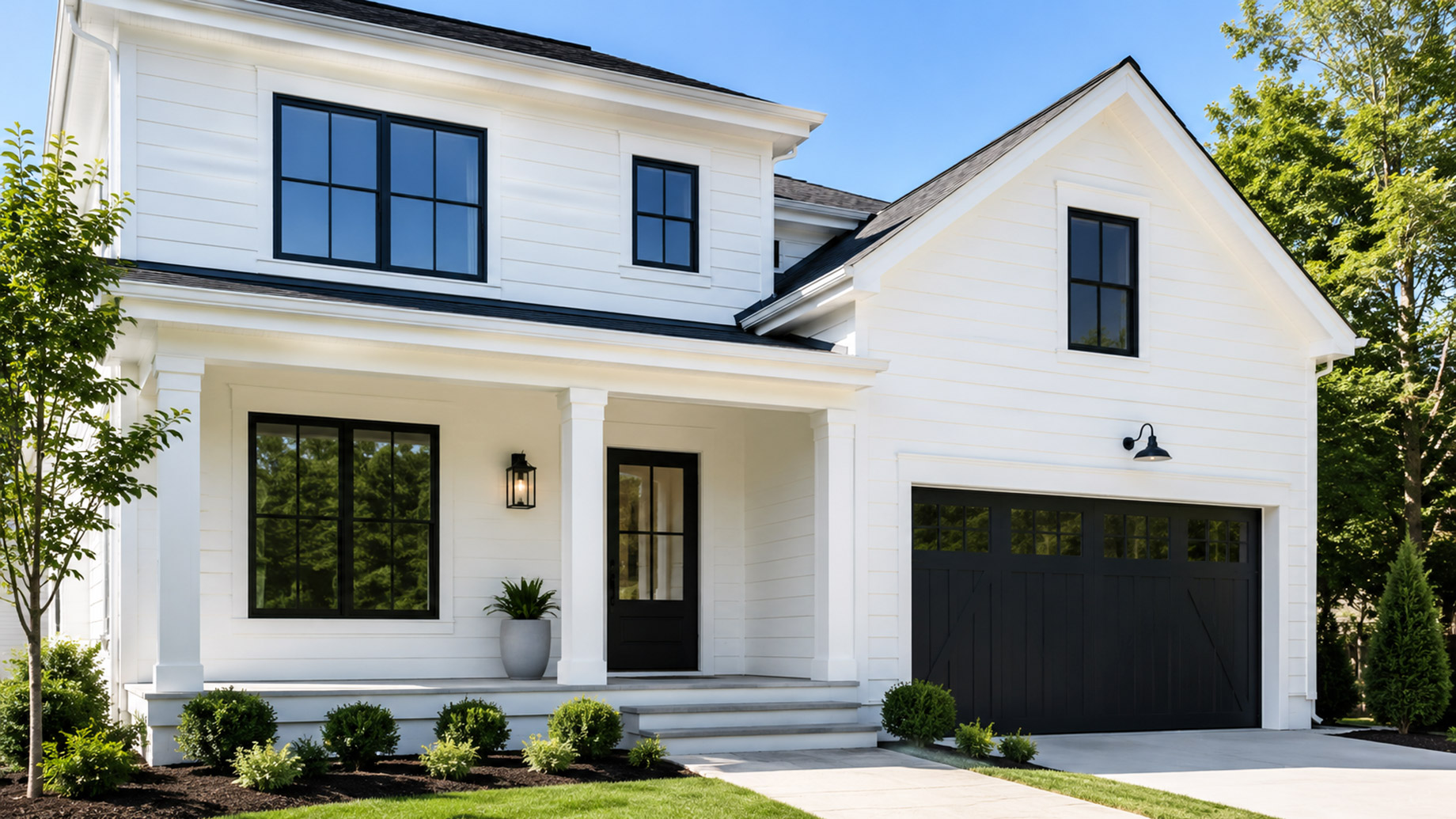 Modern white luxury house with black-framed windows, a black garage door, front porch, and landscaped yard in bright daylight.