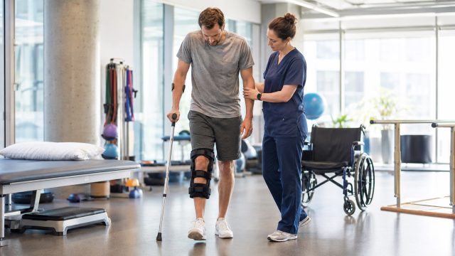 Injured man wearing a leg brace and using a cane while a physical therapist assists him during rehabilitation in a modern clinic.