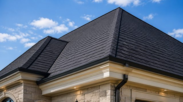 Low-angle view of a luxury stone home with a pristine dark slate-style roof, clean ridge lines, black gutters, cream trim, arched windows, and warm exterior lighting.