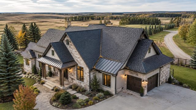 A luxury rural home with a dark shingled roof, metal roof accents, gutters, and surrounding acreage landscape near open fields and trees.