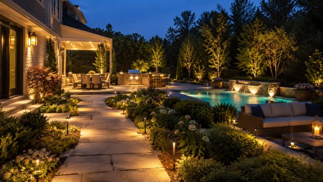 Luxury backyard with warm pathway lights, an illuminated pool, outdoor kitchen, patio dining area, and landscaped gardens at dusk.