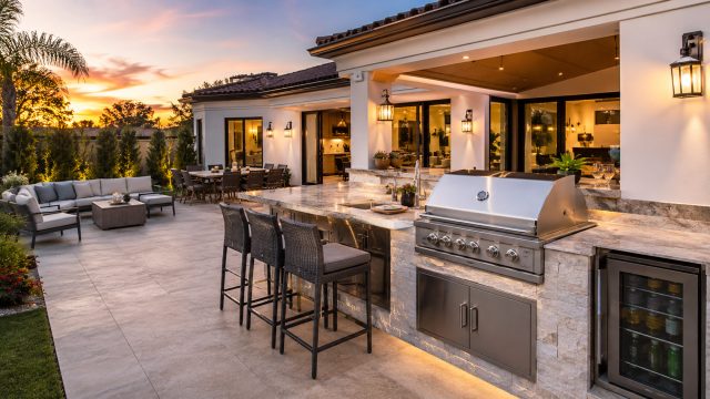 Luxury outdoor kitchen with built-in grill, stone countertops, bar seating, dining area, lounge space, and warm evening lighting.
