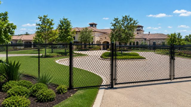 Luxury estate with black chain link fencing, gated driveway, manicured landscaping, and Mediterranean-style architecture under a sunny blue sky.