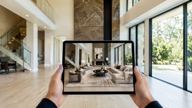 Hands hold a tablet in an empty luxury living room, showing an augmented reality view of the same space fully staged with elegant furniture, rugs, lighting, and décor.